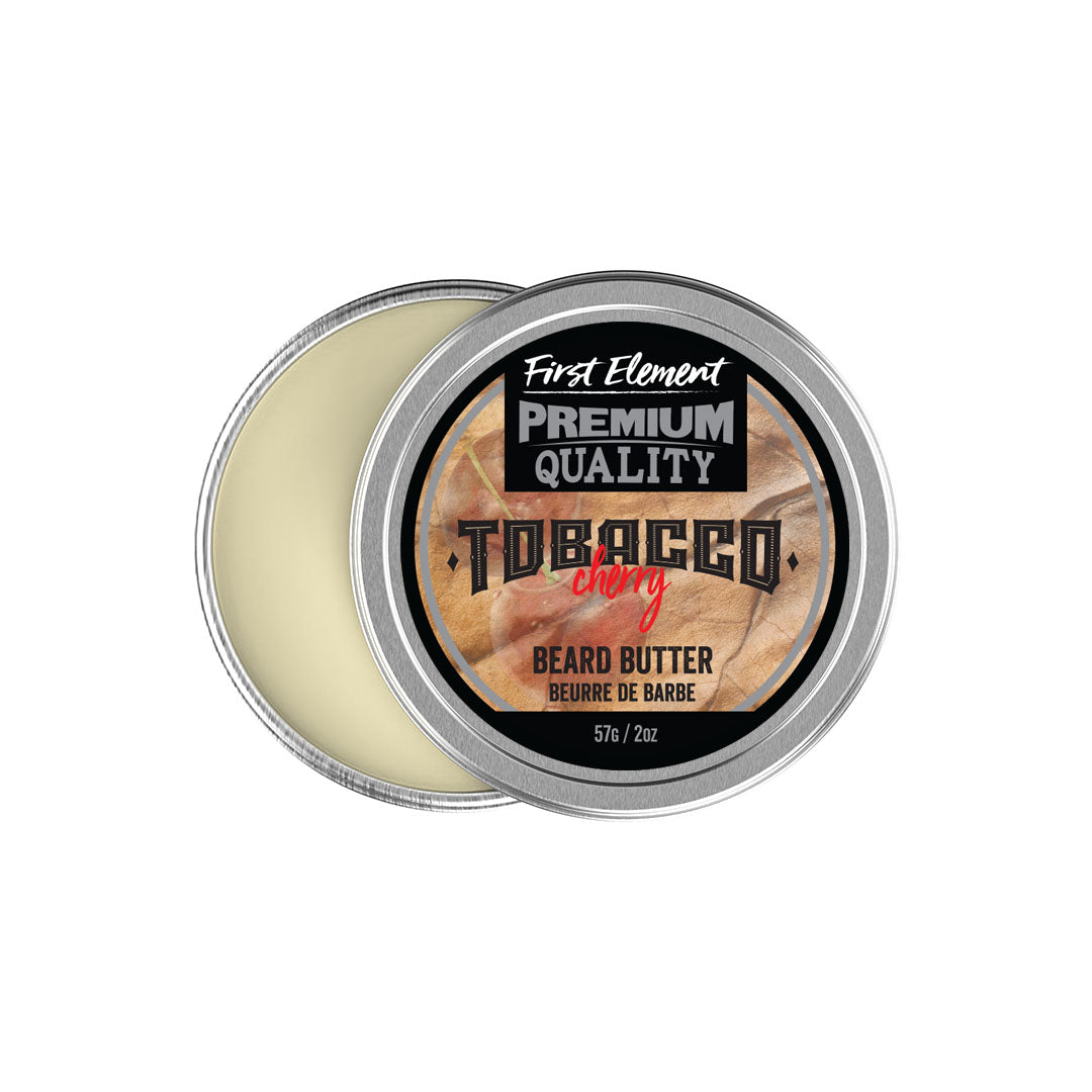 Image: A silver metal tin with a screw-on lid and tamper-evident seal, containing Cherry Tobacco scented Beard Butter. Text on the label reads 'Premium Beard Butter, Made in Canada'. The tin is surrounded by green leaves, emphasizing its natural ingredients.