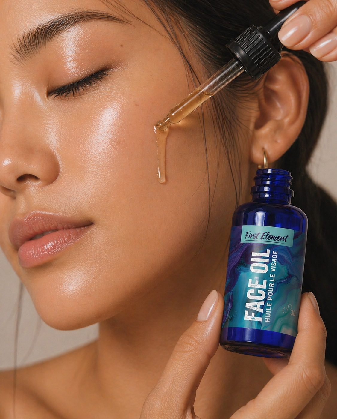 Person applying face oil from a dropper to their ear with a bottle labeled 'First Element Face Oil' nearby.