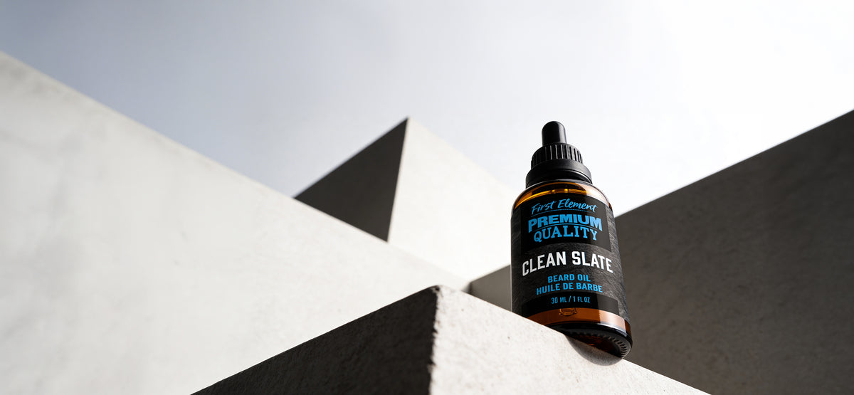 A small amber dropper bottle labeled Best Men's Premium Quality Clean Slate beard oil resting on a concrete structure against a bright sky.