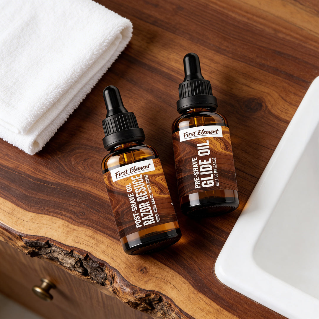 Two bottles of 'First Element' grooming products on a wooden surface with a towel and sink in the background. Pre-Shave and Post-Shave Oils.